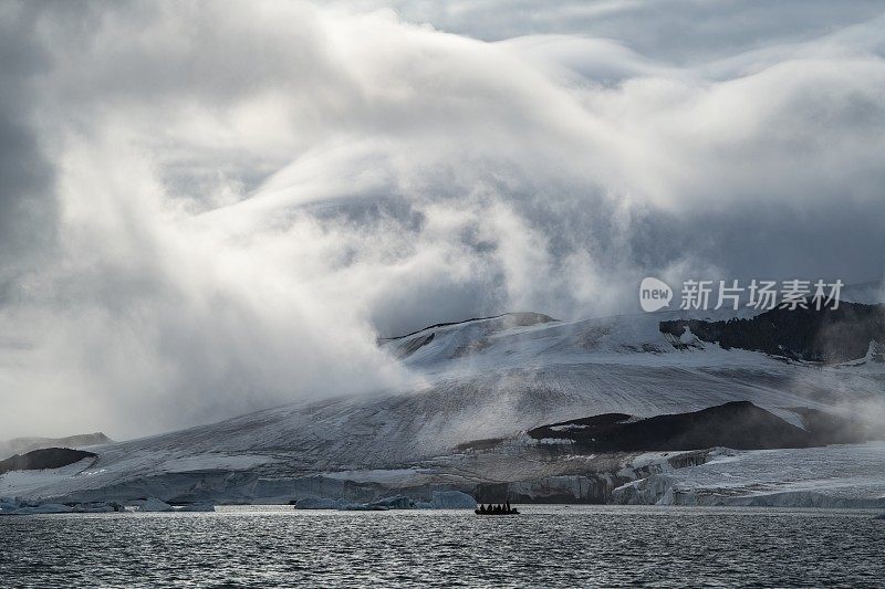 乘坐Zodiac摩托化橡皮艇的探险游轮MV Sea Spirit (Poseidon Expeditions)的乘客在冰川、出现的岩石和被太阳照亮的神秘云彩前的剪影